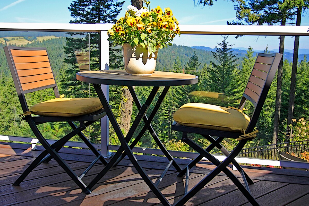 A round table set for two features yellow cushioned chairs on a deck. A potted flower arrangement adds a touch of color. The backdrop showcases a forested landscape with distant mountains and a clear blue sky.