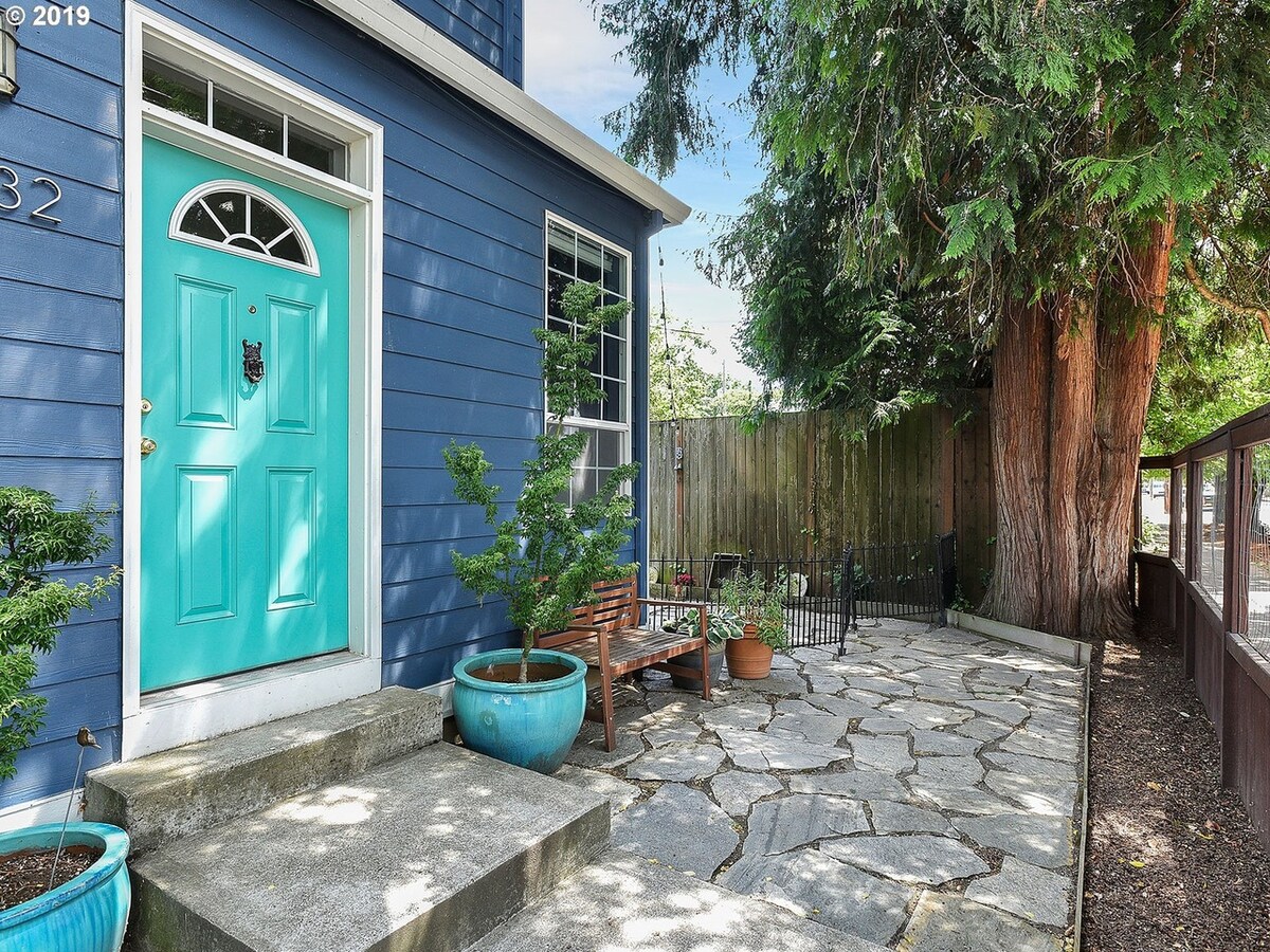 The entrance features a bright turquoise door framed by a stone walkway and planters. A seating area with wooden chairs is present, surrounded by greenery and a wooden fence, creating a welcoming outdoor space. Sunlight filters through trees, enhancing the serene environment.