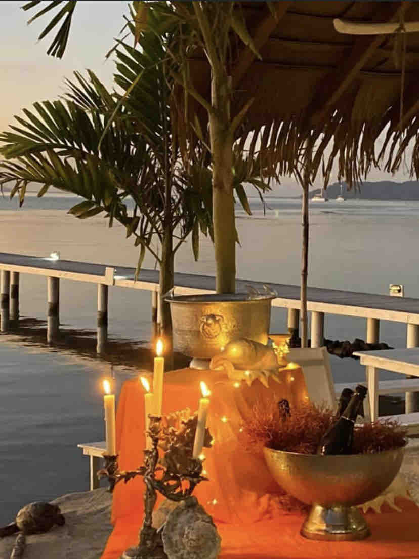 An elegantly set outdoor table features illuminated candles and a decorative bowl, with a tranquil waterfront view in the background. Palm leaves provide a natural touch, while soft light reflects off the water, enhancing the serene atmosphere during sunset.