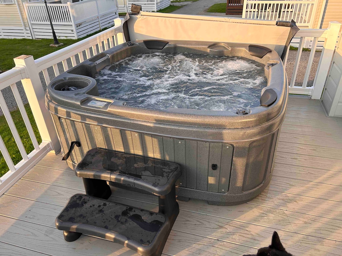 A private hot tub is situated on a wooden deck, featuring a textured exterior and bubbling water. Steps lead up to the tub, which is enclosed by a safety rail and provides an inviting space for relaxation.