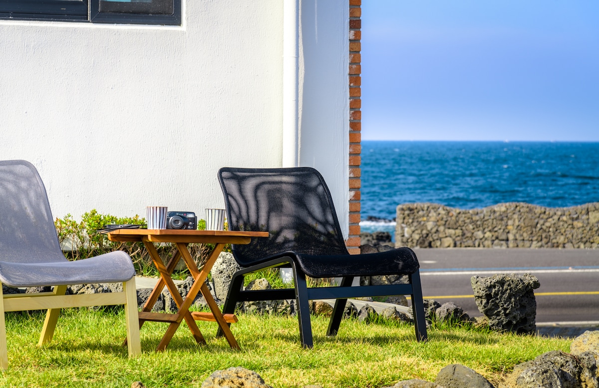 Two lounge chairs in neutral tones are positioned on a grassy area, accompanied by a small wooden table. The view features the calm ocean, creating a relaxed coastal atmosphere. A stone wall runs parallel to the nearby road, adding to the tranquil outdoor setting.