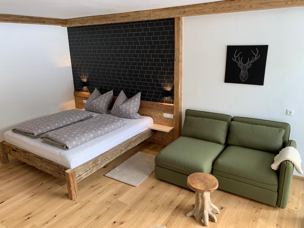 A modern alpine-style bedroom features a spacious bed adorned with decorative pillows against a dark accent wall. A green seating area with a wooden side table complements the natural wooden elements in the room, creating a cozy and inviting space.