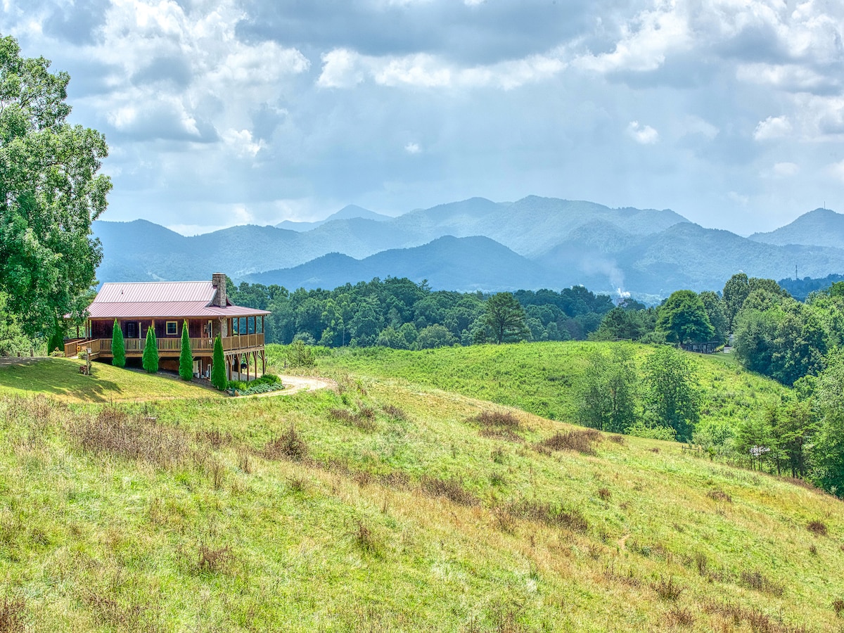 The cabin is positioned on a grassy hillside, overlooking a vast expanse of rolling mountains under a cloudy sky. The wraparound porch offers a panoramic view, showcasing the natural beauty of the surrounding landscape.