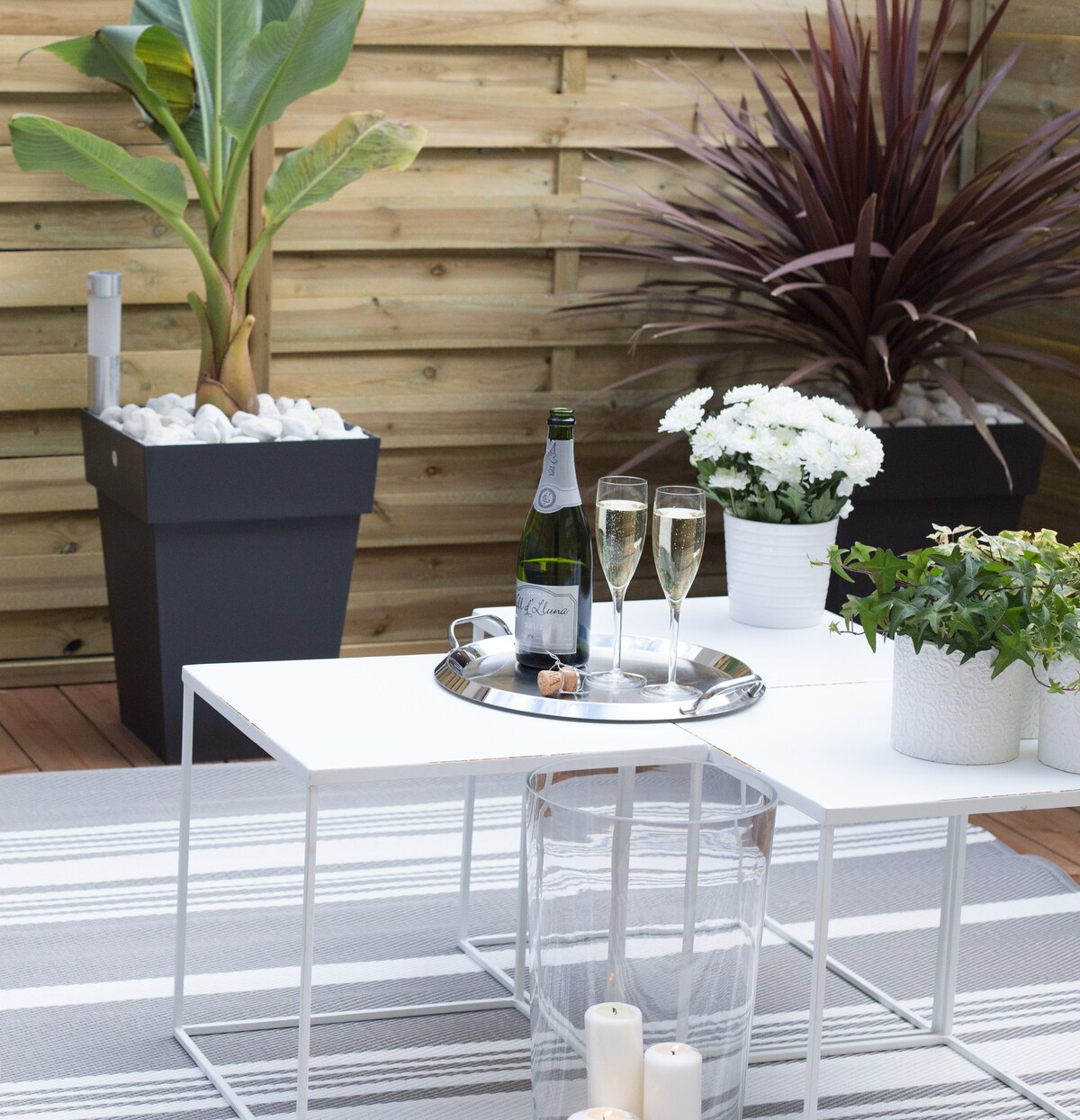 A stylish outdoor terrace is showcased, featuring a sleek white table with a silver tray holding two flutes of champagne. Potted plants, including vibrant greenery and white flowers, enhance the space. A striped rug adds texture under a setting of soft candlelight.