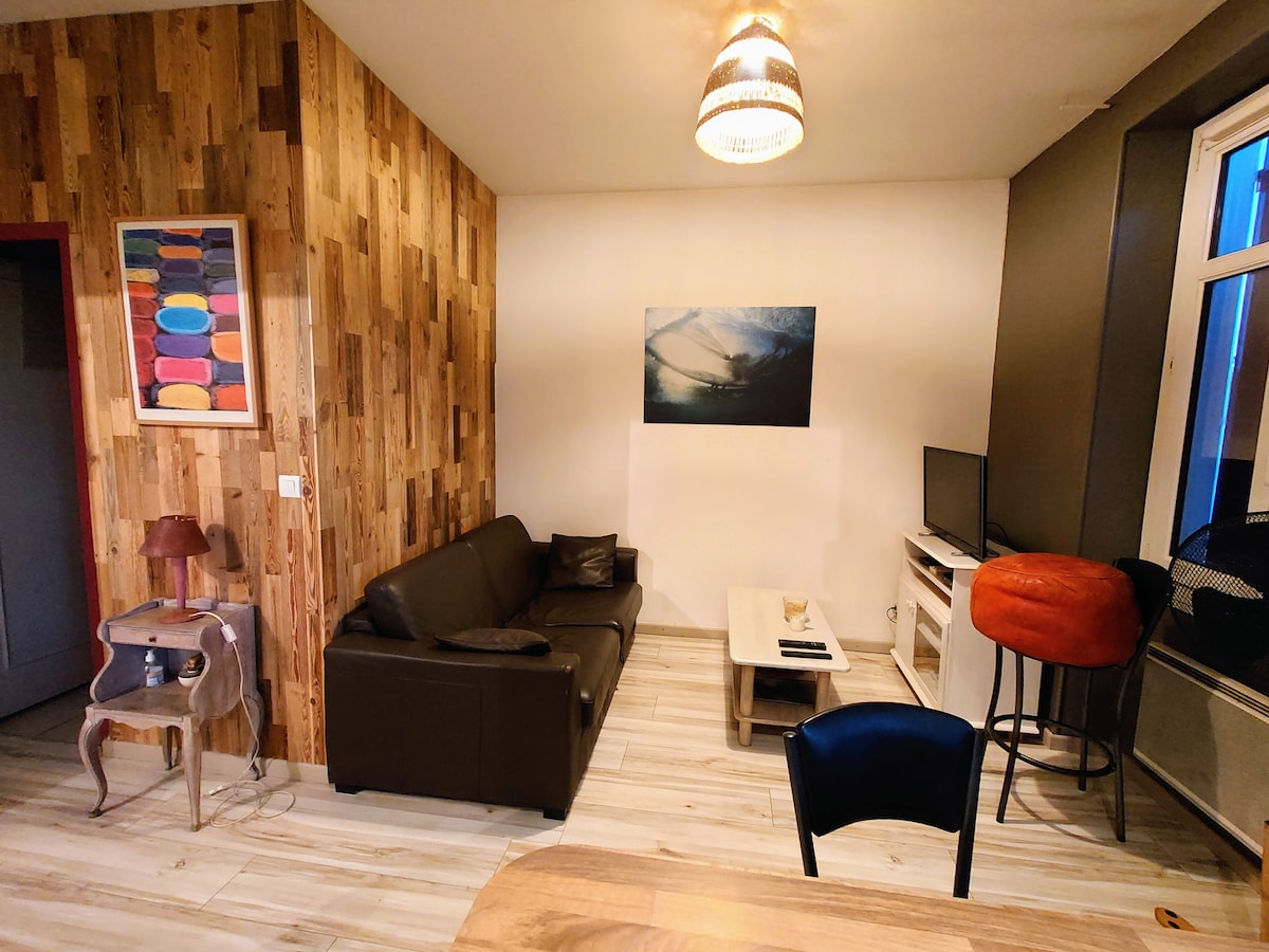 A cozy living area is presented, featuring a dark sofa and a light coffee table. A television sits on a stand, while a wooden accent wall adds texture. Light from a nearby window illuminates the space, complemented by a unique pendant light overhead.