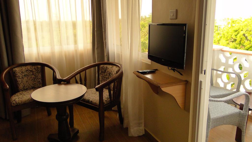 A seating area features two wooden chairs with patterned upholstery, positioned around a small circular table. Light-colored curtains frame the window, allowing natural light to fill the space. A wall-mounted television is visible, along with a balcony view that can be accessed from the room.