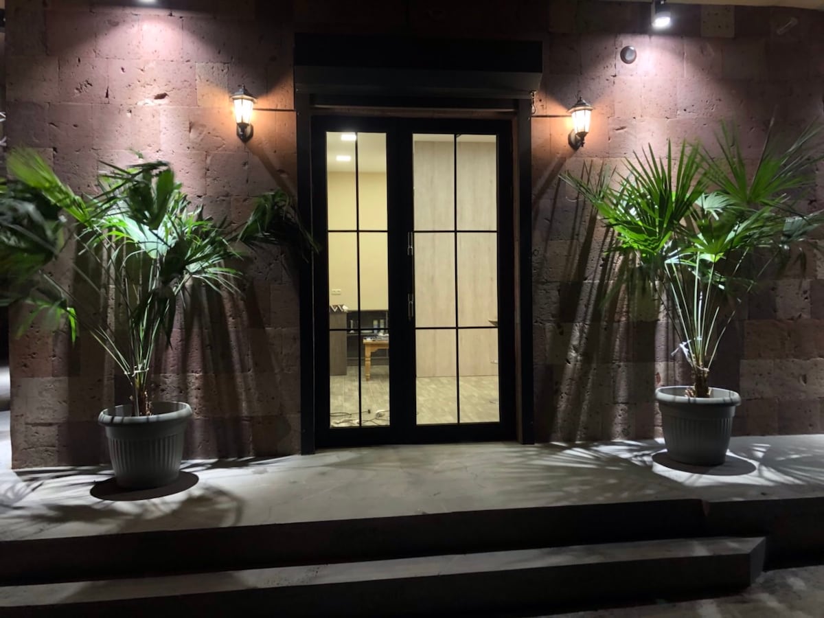 The entrance is framed by two tall potted plants, providing a welcoming feel. A set of glass doors leads into the interior, flanked by warm lighting fixtures that highlight the textured stone wall. The steps are wide, facilitating easy access.