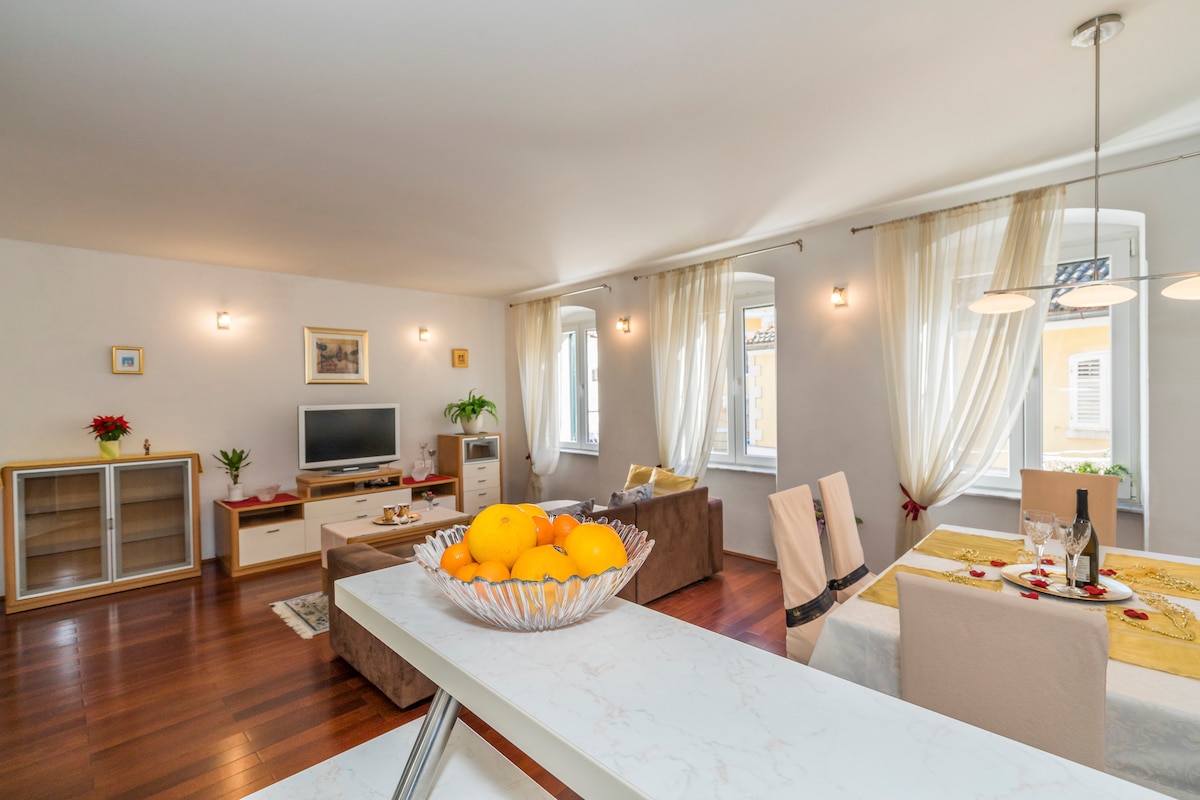 A bright and airy living space features large windows allowing natural light to fill the room. A dining area is set with a table, food, and wine, while a cozy seating area includes a television. Light curtains flutter gently in the breeze.