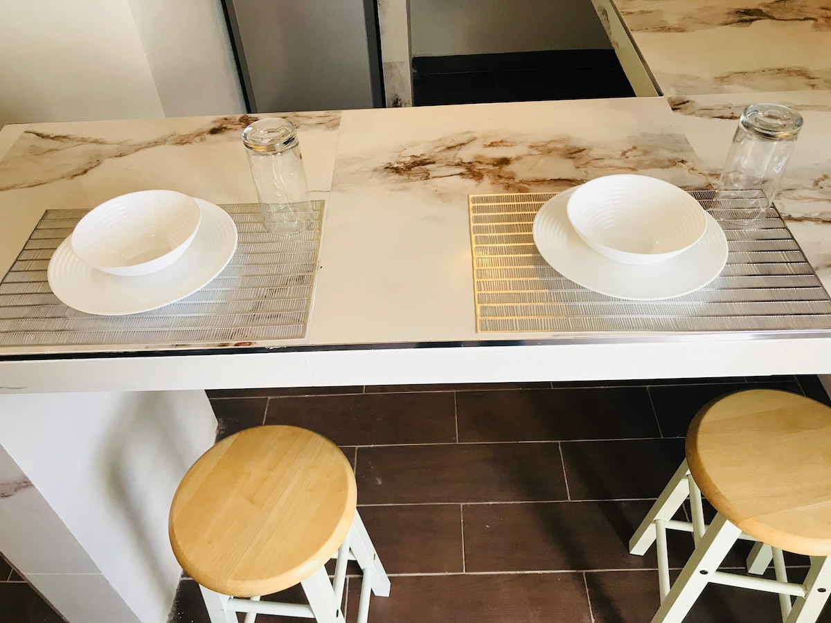 A dining area features a clean, marble-top table set for two. Each place setting includes a bowl placed on a silver mat, accompanied by a glass. Two wooden stools with light finishes are positioned beneath the table.