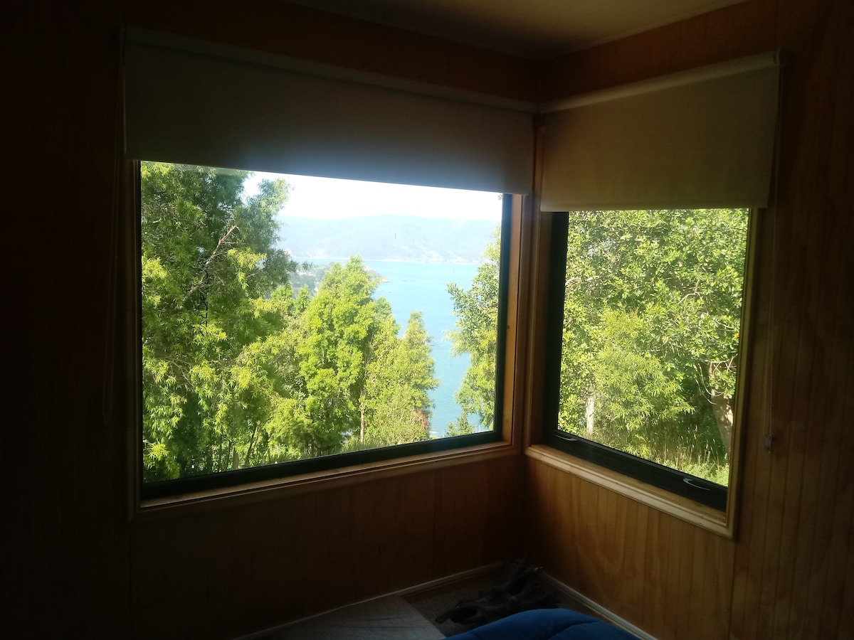 Large windows frame a scenic view of the water and surrounding greenery. Natural light enters the comfortable interior, highlighting wooden walls and a simple sleeping area. Shades are partially drawn, allowing for a peaceful atmosphere while observing the landscape beyond.