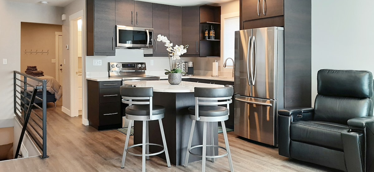 A modern kitchen space is showcased with sleek, dark cabinetry and stainless steel appliances. An island with seating for two is centered, complemented by a reclining chair nearby. Soft lighting enhances the overall space, creating a functional cooking and dining area.