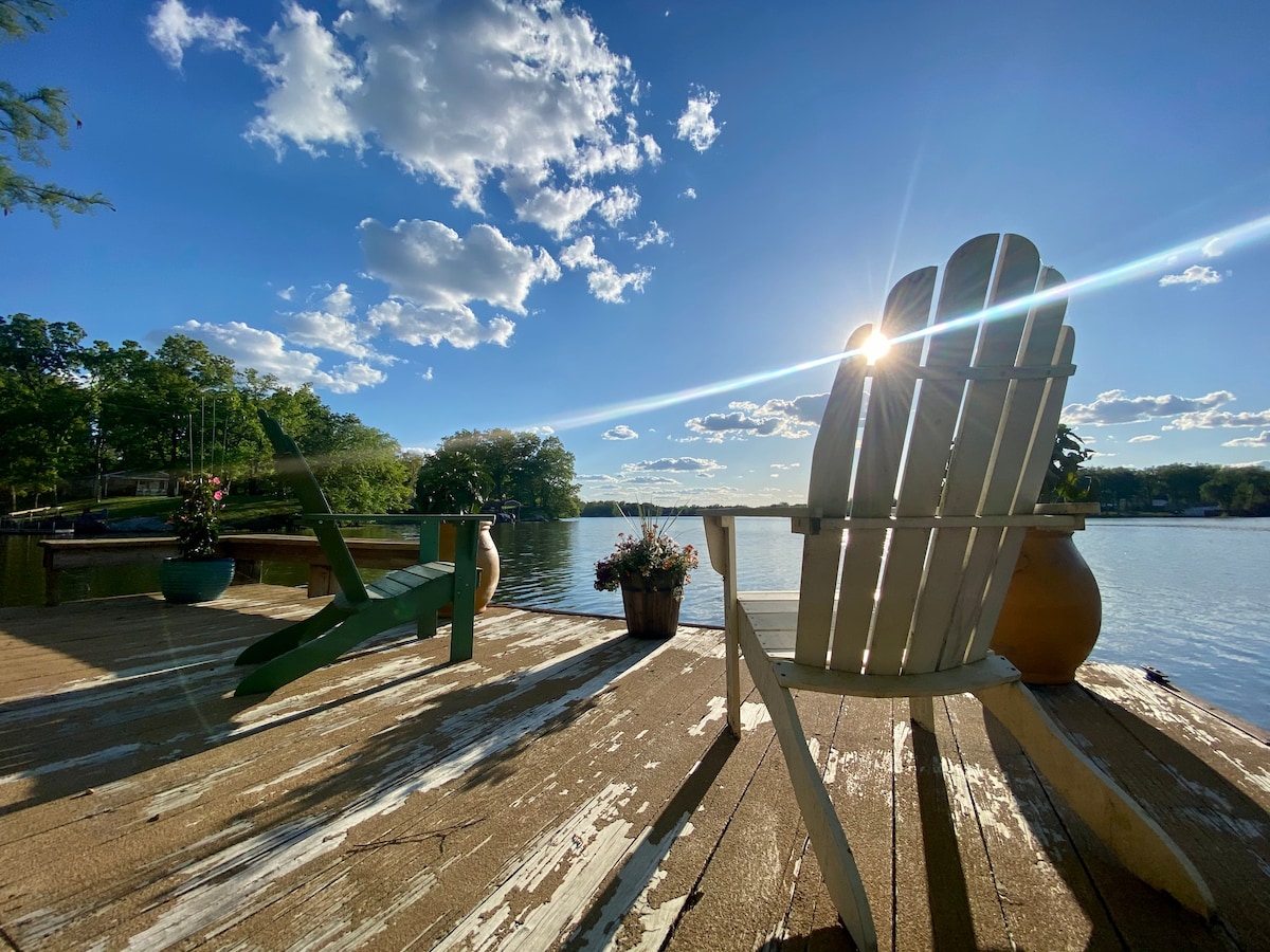 A serene lakeside view is captured from a wooden deck, featuring two chairs facing the water. A flower pot and decorative vase are positioned nearby, while the sunlight reflects off the lake's surface. Lush green trees line the opposite shore, enhancing the tranquil scene.