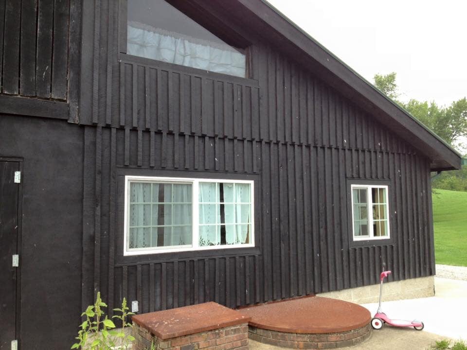 The exterior of a black lodge features a combination of wooden paneling and large windows. A small, round stone patio area is present in front, complemented by a pink child’s scooter parked nearby. Lush green countryside can be seen in the background.