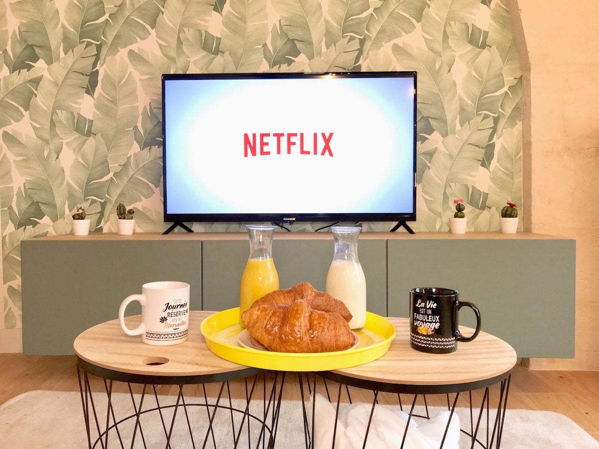 A television displaying the Netflix logo is positioned on a minimalist cabinet against a leafy wallpaper. A yellow tray with a croissant is placed on a low coffee table alongside cups containing orange juice and milk. Small potted plants add a touch of greenery.