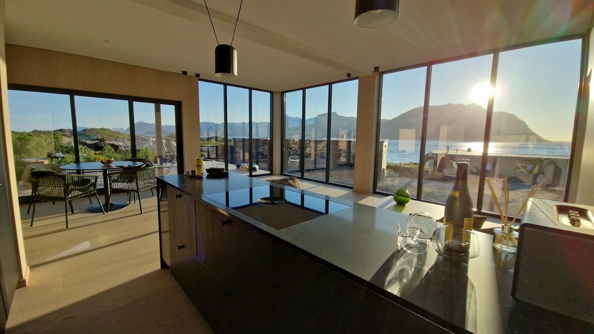 A modern kitchen is featured with large windows allowing for ample natural light and views of the surrounding landscape. Black stone countertops contrast with light-colored cabinetry. A dining table with chairs is visible outside, framed by the scenic backdrop of mountains and the ocean.