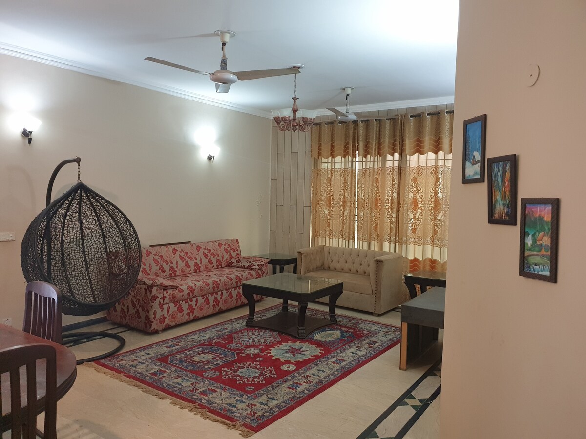 A spacious lounge area is designed with a blend of seating options, including a patterned sofa and a hanging chair. Natural light filters through sheer curtains, illuminating the decorative rug on the floor. Wall art adds a touch of color to the neutral-toned walls.