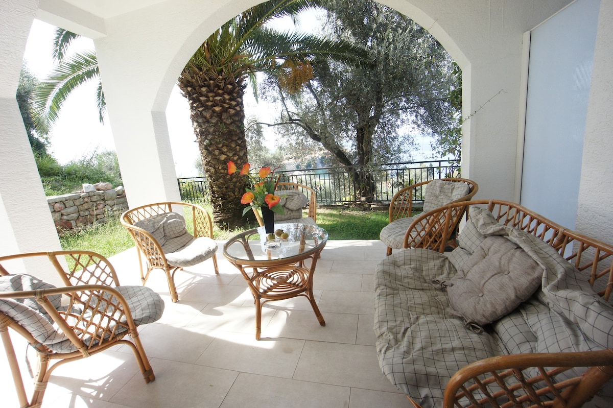 A shaded outdoor seating area is featured, furnished with four rattan chairs and a glass table. A cluster of vibrant flowers decorates the table. Lush greenery and a palm tree are visible in the background, enhancing the tranquil setting.