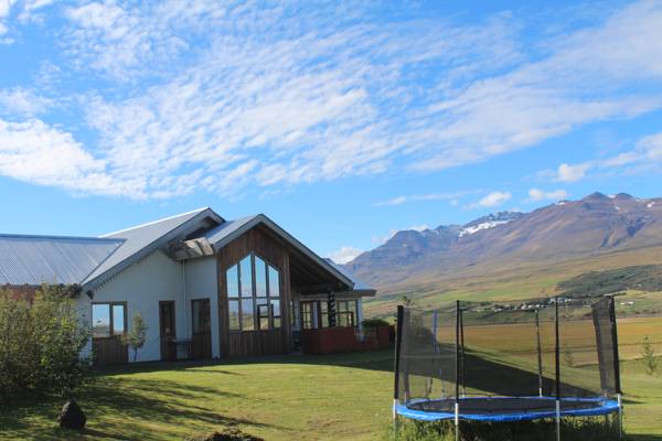 The image displays a modern house with large windows reflecting the surrounding landscape. A trampoline is positioned in the foreground, while rolling hills and mountains can be seen in the background under a clear blue sky.