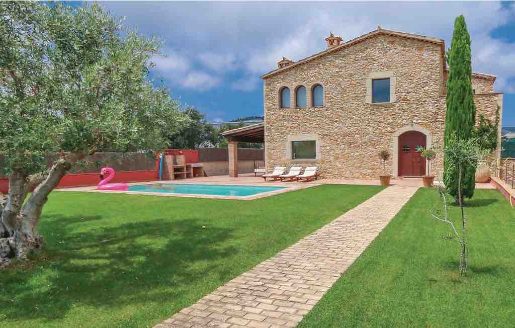 A stone house features large arched windows and a welcoming front door. A neatly paved path leads through the green lawn to a private pool surrounded by sun loungers. An olive tree and a tall cypress add to the outdoor space.