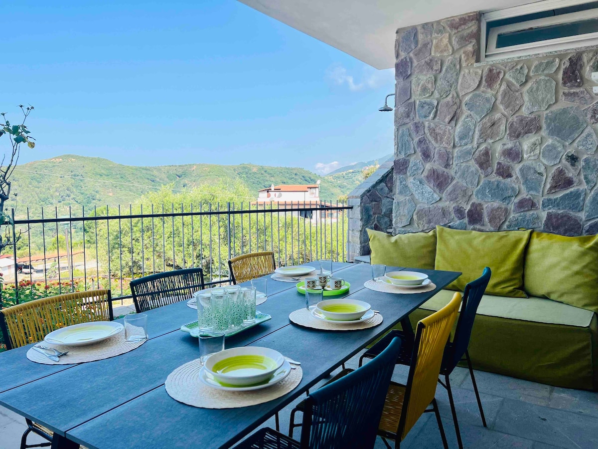 An outdoor terrace features a large dining table set for six, accompanied by colorful chairs. A comfortable seating area is presented with soft cushions. Rolling green hills are visible in the background under a clear blue sky, enhancing the tranquil ambiance of the space.