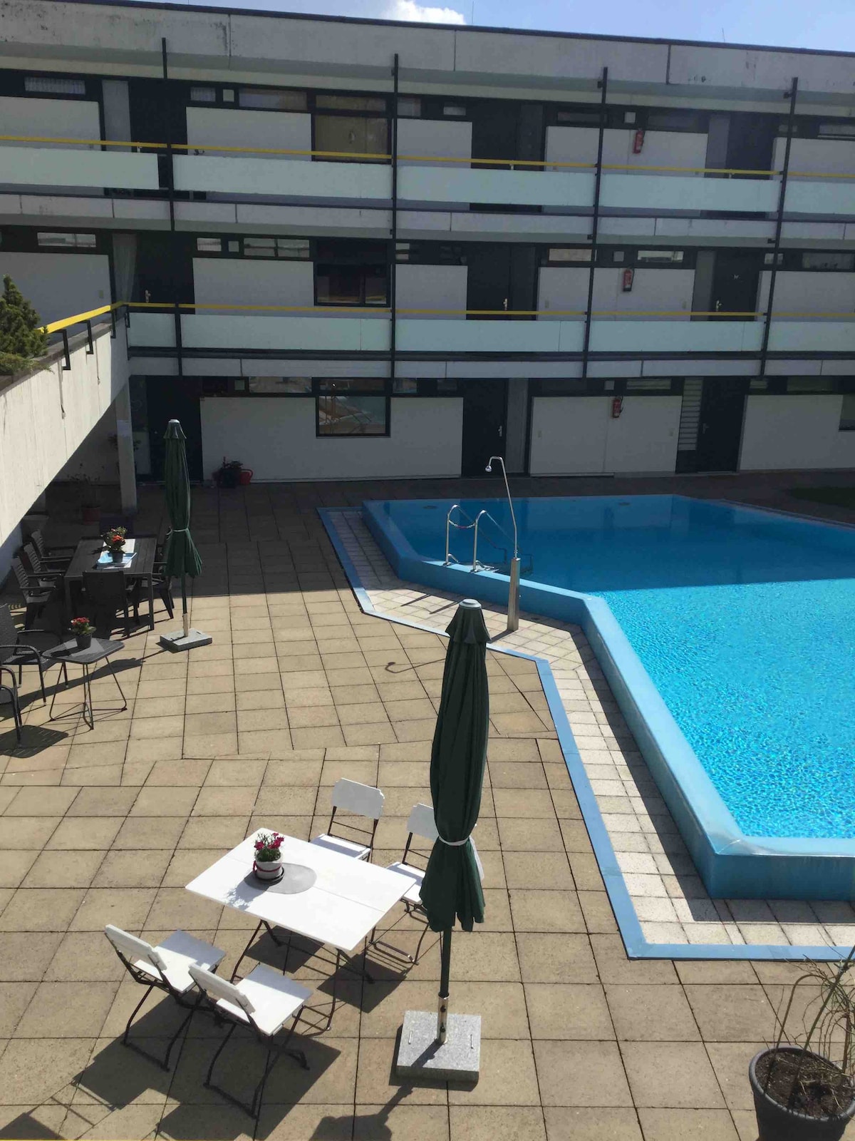 An outdoor pool is visible, surrounded by a tiled patio. Several chairs with green umbrellas are arranged near a dining table. The building features multiple balconies and windows on either side of the pool area, creating a communal space for relaxation.