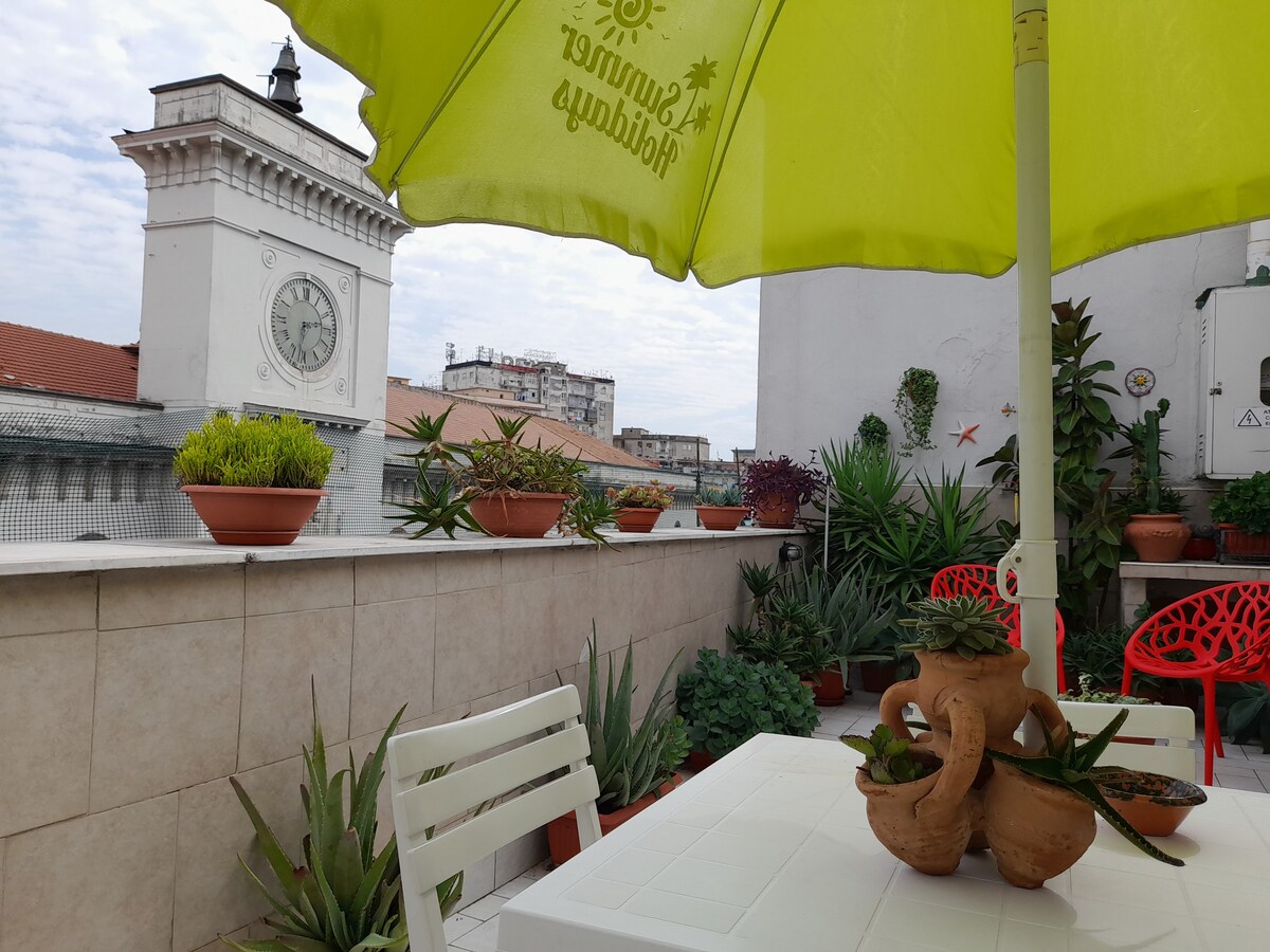 A terrace is depicted, featuring a white table and chairs under a large green umbrella. Various potted plants are arranged around the space. In the background, a clock tower adds a scenic element to the view.