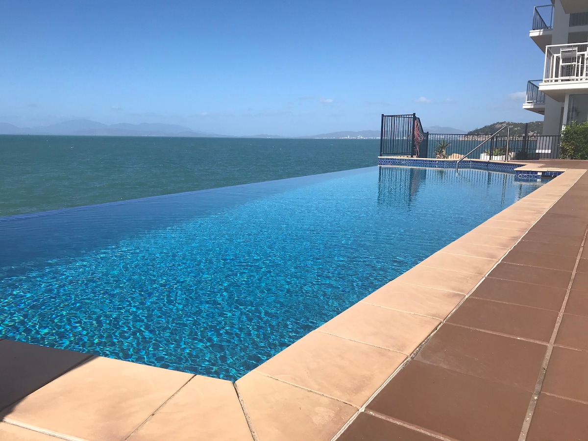 An infinity pool extends toward the horizon, reflecting shades of blue from the water and sky. Mountains are visible in the distance, while the pool's edge seamlessly meets the sea. Accessible lounging areas are available along the poolside, enhancing the relaxed environment.