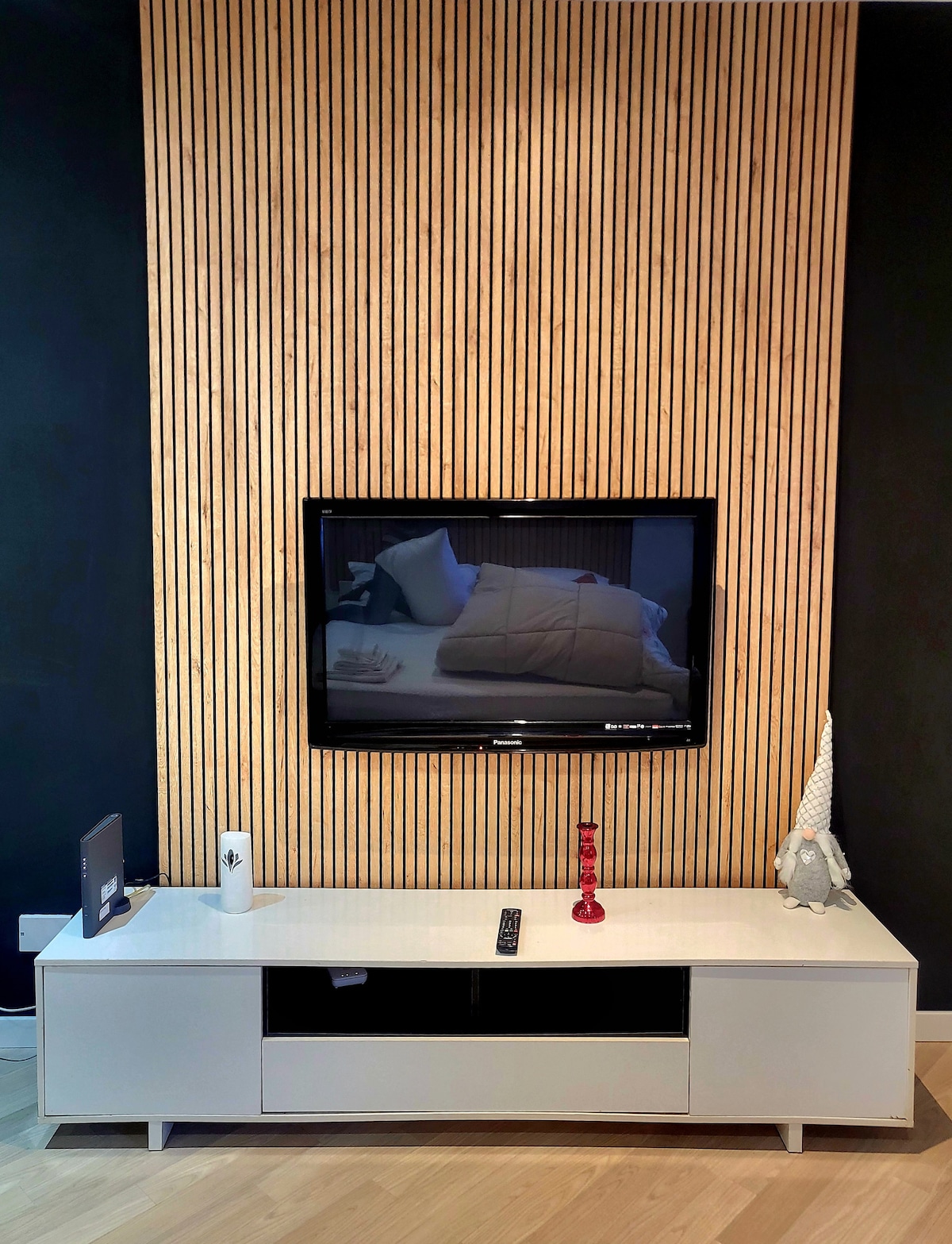 A modern entertainment area features a wall-mounted television above a sleek, white media console. Natural wood paneling creates a warm backdrop, while a decorative object and a remote control are positioned on the console. Soft lighting enhances the minimalist design.