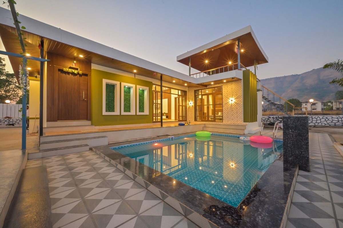 A modern exterior view of the property showcases a private swimming pool surrounded by a well-designed patio area. Floor-to-ceiling windows invite natural light into the house, with vibrant green accents and a mountain backdrop enhancing the serene setting.