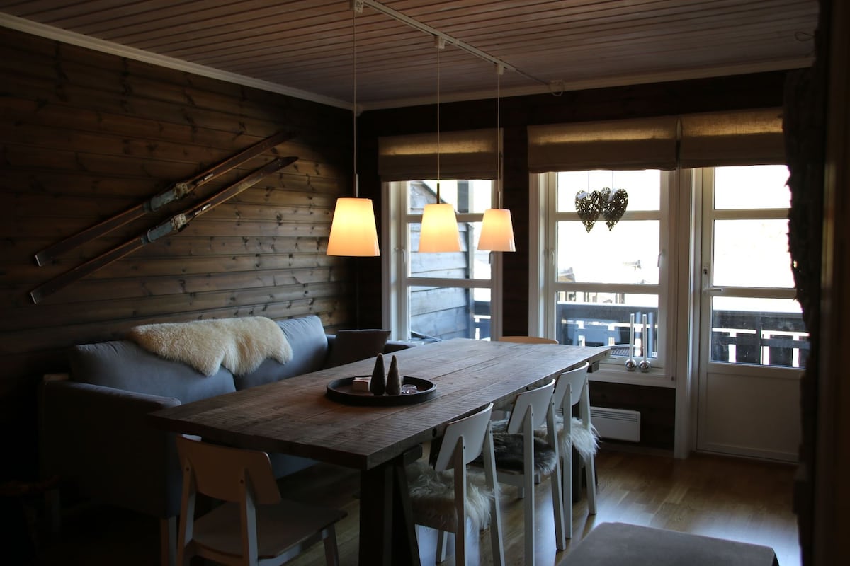 A dining area is featured with a large wooden table surrounded by white chairs. Three pendant lights hang above, casting a warm glow. A cozy sofa is positioned against a wooden wall adorned with vintage ski equipment, and large windows offer glimpses of the surrounding landscape.