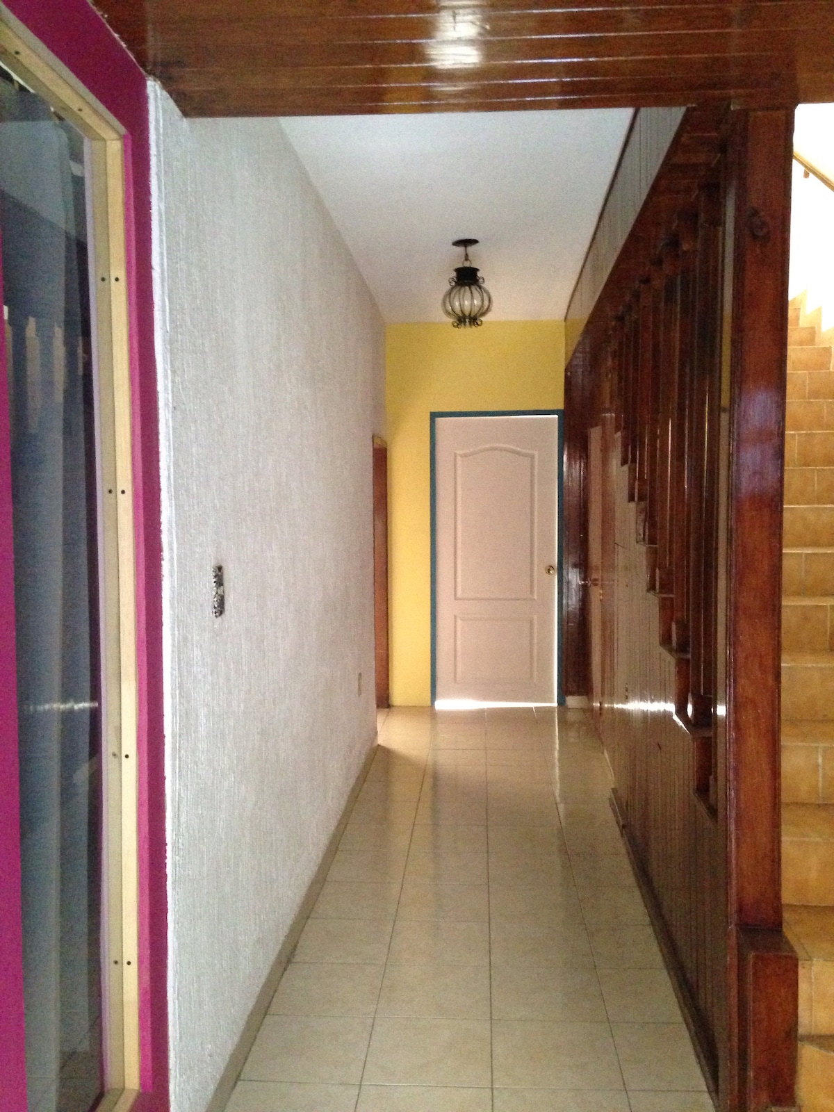 A narrow hallway is viewed, showcasing smooth tile flooring and a combination of light-colored walls with a vibrant pink accent. A staircase is visible at the end, bordered by wooden banisters. Natural light filters in from the doorway, illuminating the space.