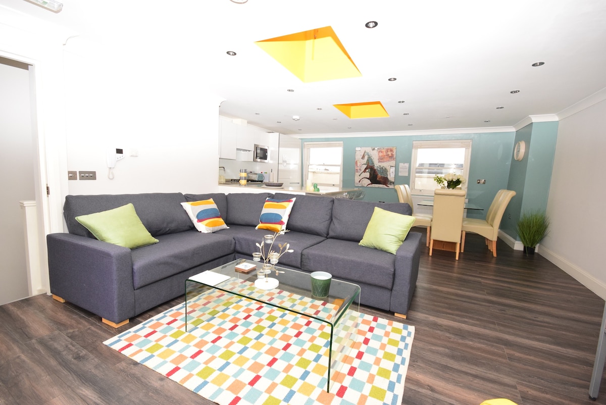 A spacious living area is presented, featuring a large sofa adorned with cushions in various colors. A colorful patterned rug covers the floor under a glass coffee table. The open-plan layout shows a dining area in the background, accompanied by natural light from nearby windows.