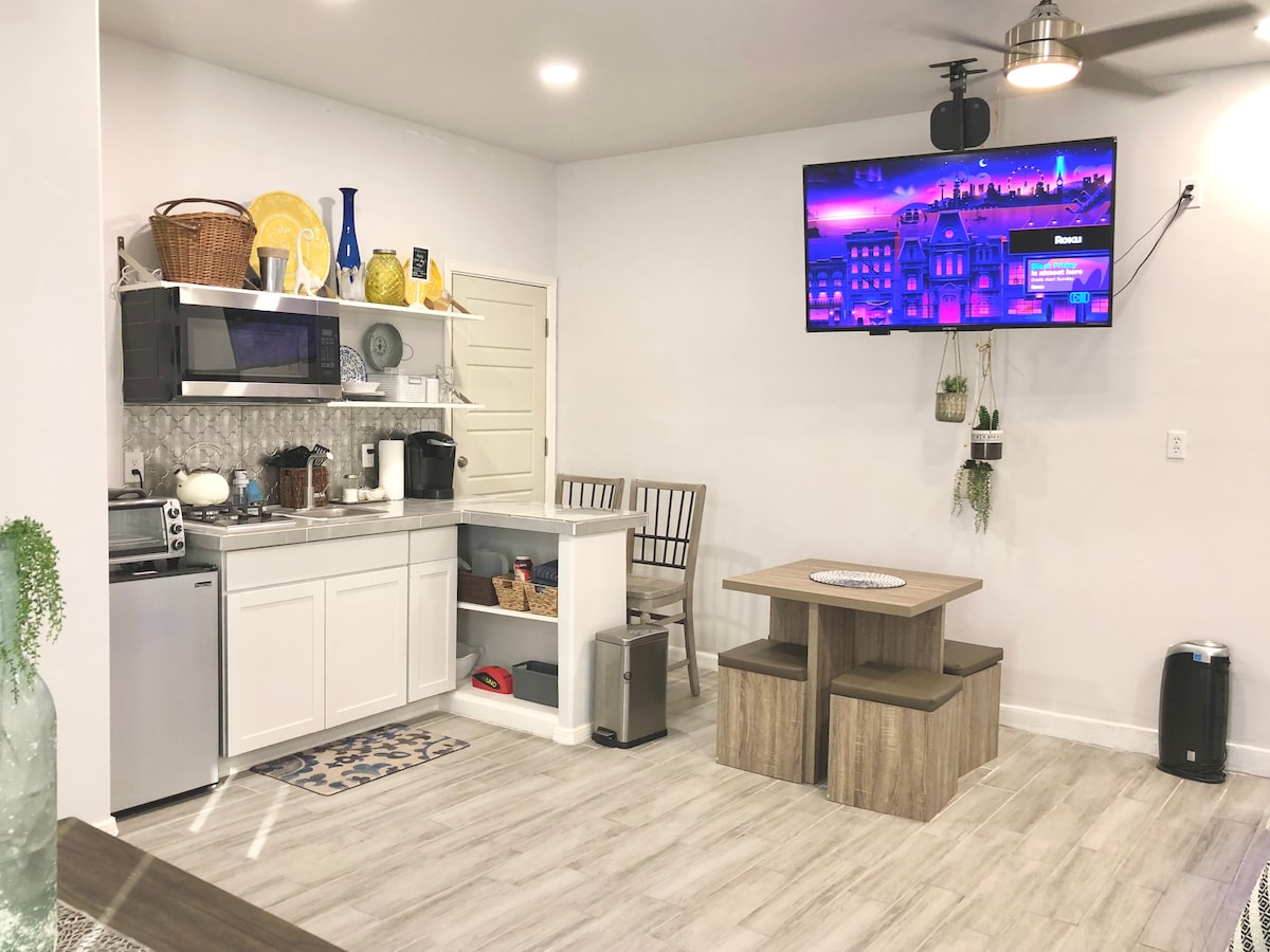 The kitchenette is equipped with a compact fridge, microwave, and stove, accentuated by shelving displaying decorative items. A modern television is mounted on the wall. A dining table and four chairs are positioned nearby, creating a functional and inviting space.