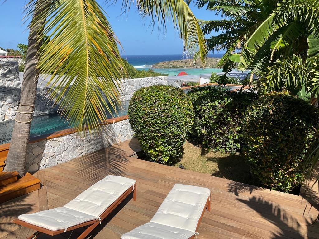 A sunlit outdoor space features two lounge chairs positioned on a wooden deck, surrounded by lush greenery. The view includes a glimpse of the ocean and a landscaped area, enhancing the inviting atmosphere of relaxation.