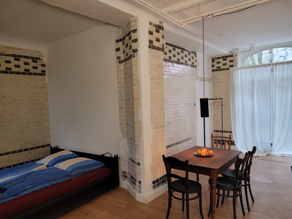 A spacious room includes a cozy sleeping area with a bed dressed in blue and white bedding. A wooden dining table is surrounded by four chairs. Textured wall tiles add character, while large windows draped with sheer curtains allow natural light to fill the space.