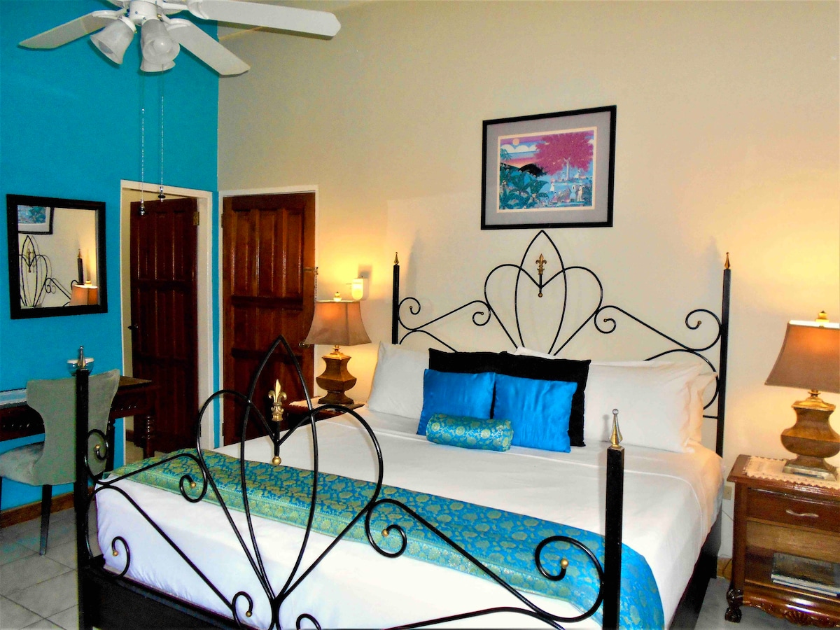 A spacious room is highlighted by a king-sized bed with a decorative blue and green bed runner. Elegant wall art and a large mirror enhance the decor. A cozy seating area is visible, complemented by tasteful lamps and a ceiling fan, creating a comfortable ambiance.