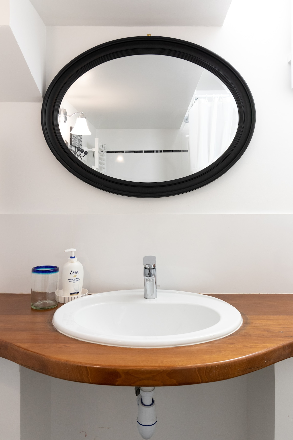 A clean and modern bathroom sink area is displayed, featuring a white circular basin set on a wooden countertop. A round black-framed mirror reflects the space above. A soap dispenser and a glass are positioned nearby, contributing to the neat appearance.