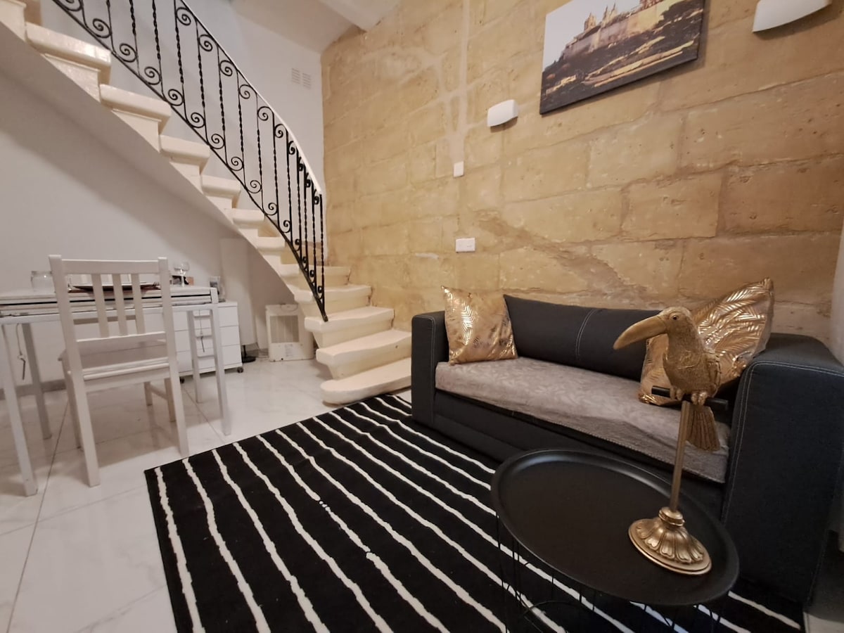 A modern living area features a black and white striped rug, a dark sofa with decorative cushions, and a small round table for dining. A stylish sculpture sits on a side table, while a sleek staircase leads to an upper level, enhancing the space's openness.