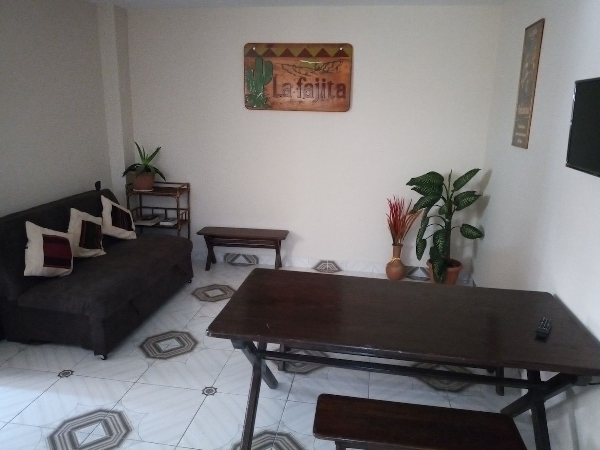 A living area is shown with a dark brown sofa adorned with cushions. A wooden table occupies the center, surrounded by a few chairs. A decorative wall sign is visible, along with potted plants in the corners, enhancing the space's inviting feel.
