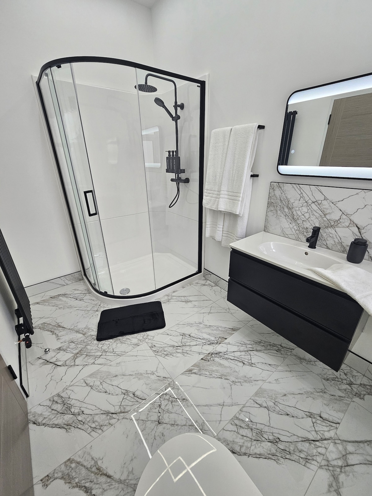 A modern bathroom showcases a sleek glass shower enclosure with a rainfall showerhead. The floor is tiled with white and gray marble patterns. A vanity with a minimalist design is equipped with a sink, complemented by a large mirror and essential toiletries on display.