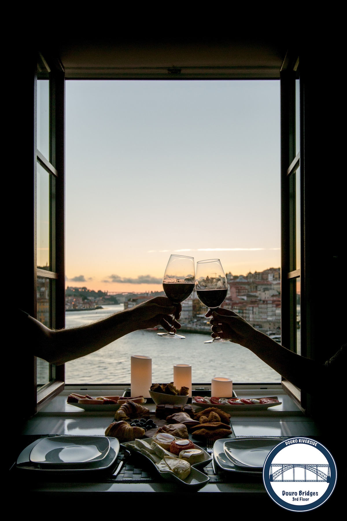 Two hands are raised in a toast with glasses of red wine against a sunset backdrop over the Douro River. A table is set with an assortment of pastries and candles, inviting guests to enjoy the scenic view and relaxing atmosphere.
