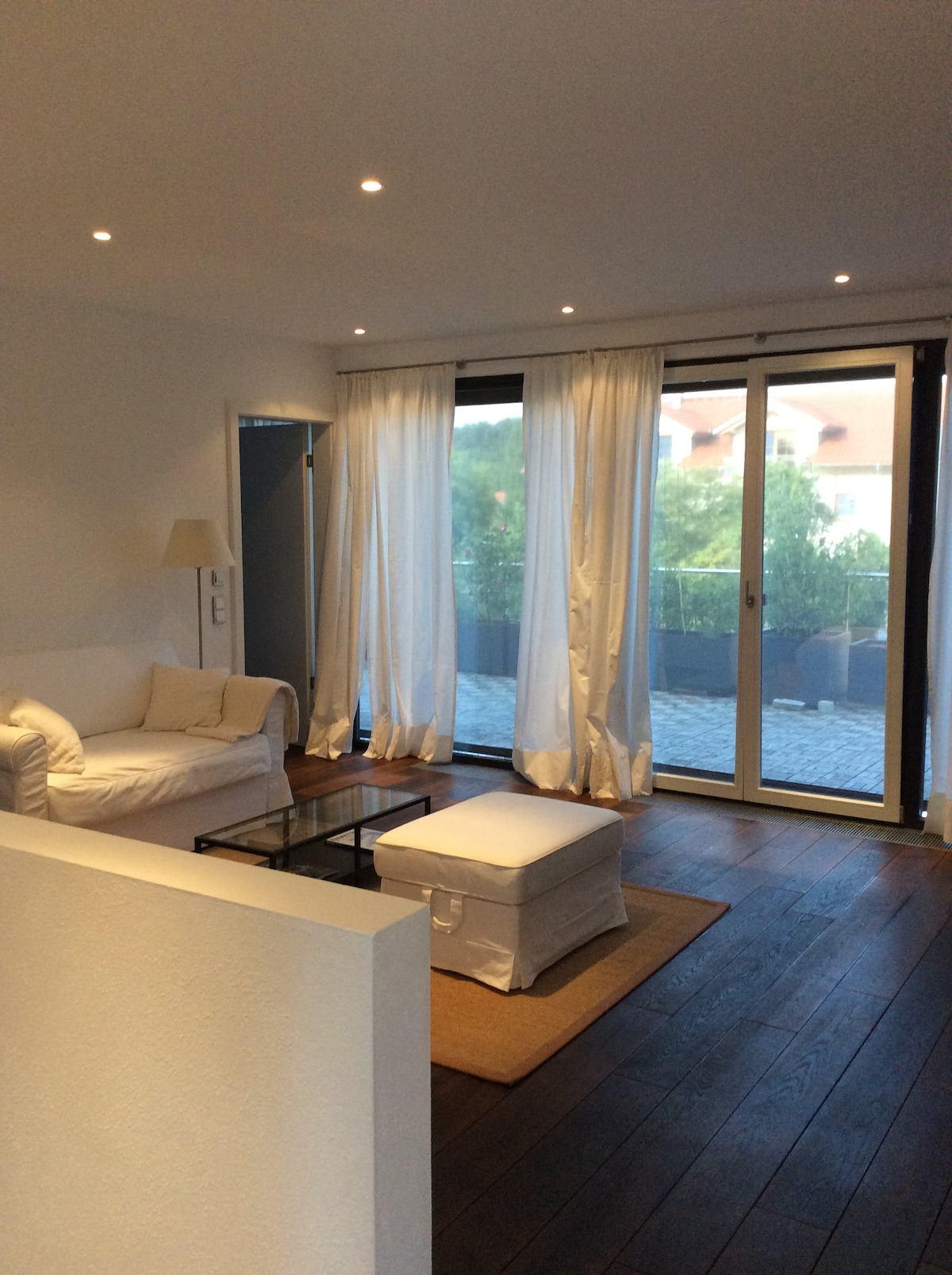 A bright living area features large glass doors that open to a balcony, allowing ample natural light to fill the space. Light, airy curtains frame the doors, while wooden flooring and a neutral color palette create a calming atmosphere. A comfortable white sofa and a glass coffee table are visible.