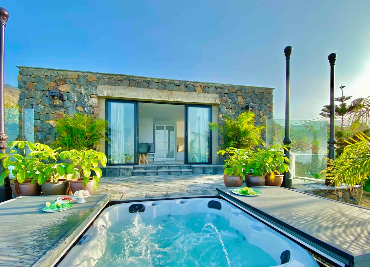 A private jacuzzi is positioned on a sunlit terrace, surrounded by lush tropical plants. Beyond, large glass doors reveal the interior space of a stone house. Bright sky and soft light enhance the serene atmosphere of this outdoor area.