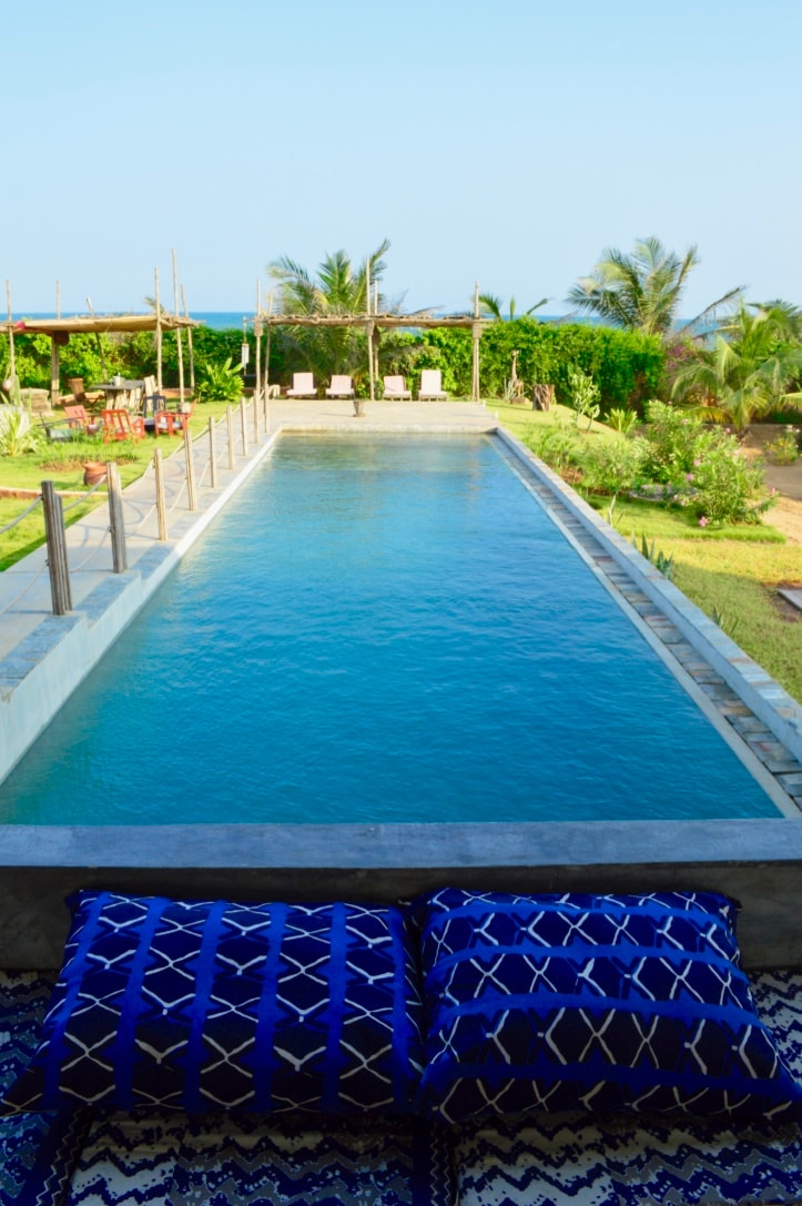 A serene swimming pool stretches into the distance, surrounded by lush greenery and palm trees. Comfortable lounges are positioned nearby, providing a perfect spot for relaxation. Vibrant cushions in blue and white patterns are placed on a woven mat, inviting guests to enjoy the peaceful outdoor space.
