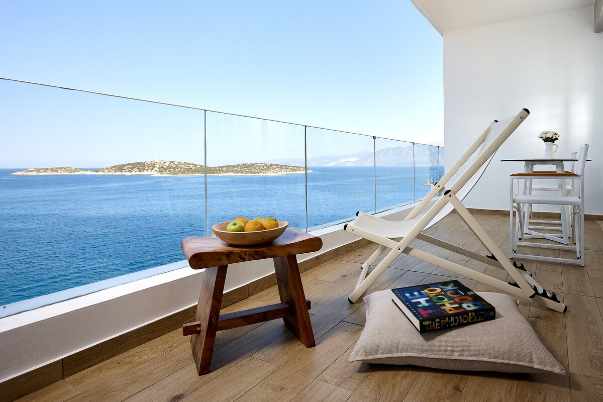 A spacious balcony offers unobstructed views of the sea and distant islands. A wooden table and two chairs provide a place to relax. A lounge chair is positioned near a small wooden table holding a bowl of citrus fruits, while a soft cushion lies on the floor.