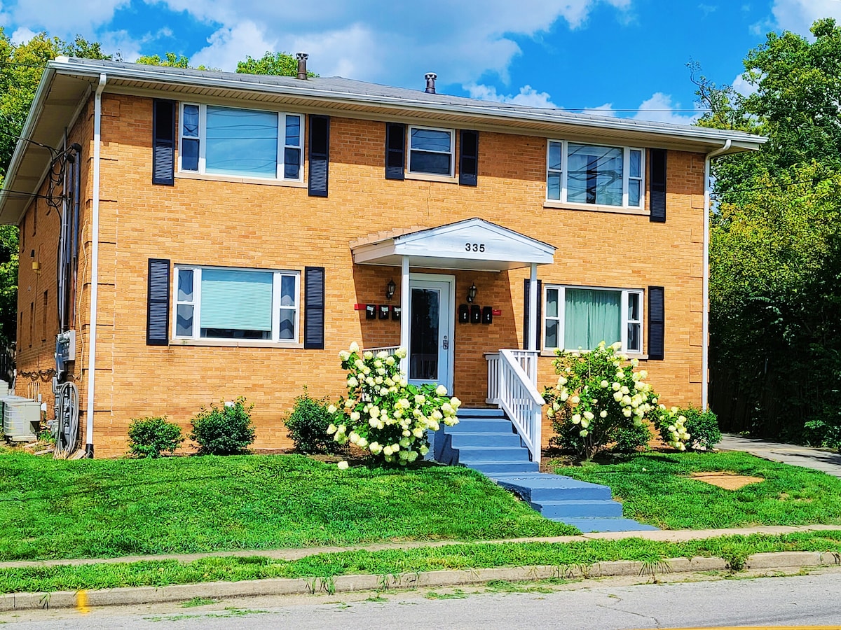 The exterior of a two-story brick apartment building is shown, featuring blue shutters and a welcoming front porch. Green grass and landscaping with blooming white flowers surround the entrance, while the sky is clear with scattered clouds.
