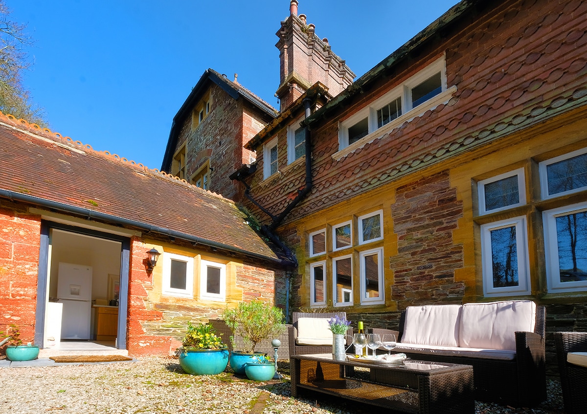The external view showcases a historic country house with textured stone and wood accents. Comfortable seating is arranged on a light stone patio, complemented by potted plants. Bright windows reflect the sunlight, enhancing the inviting exterior space.