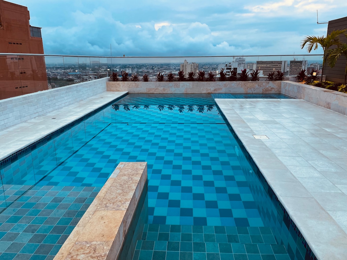 A rooftop pool is presented with clear blue waters and a checkered pattern along the border. Smooth, light-colored tiles surround the pool, and a glass railing provides views of the city skyline under a mostly cloudy sky.
