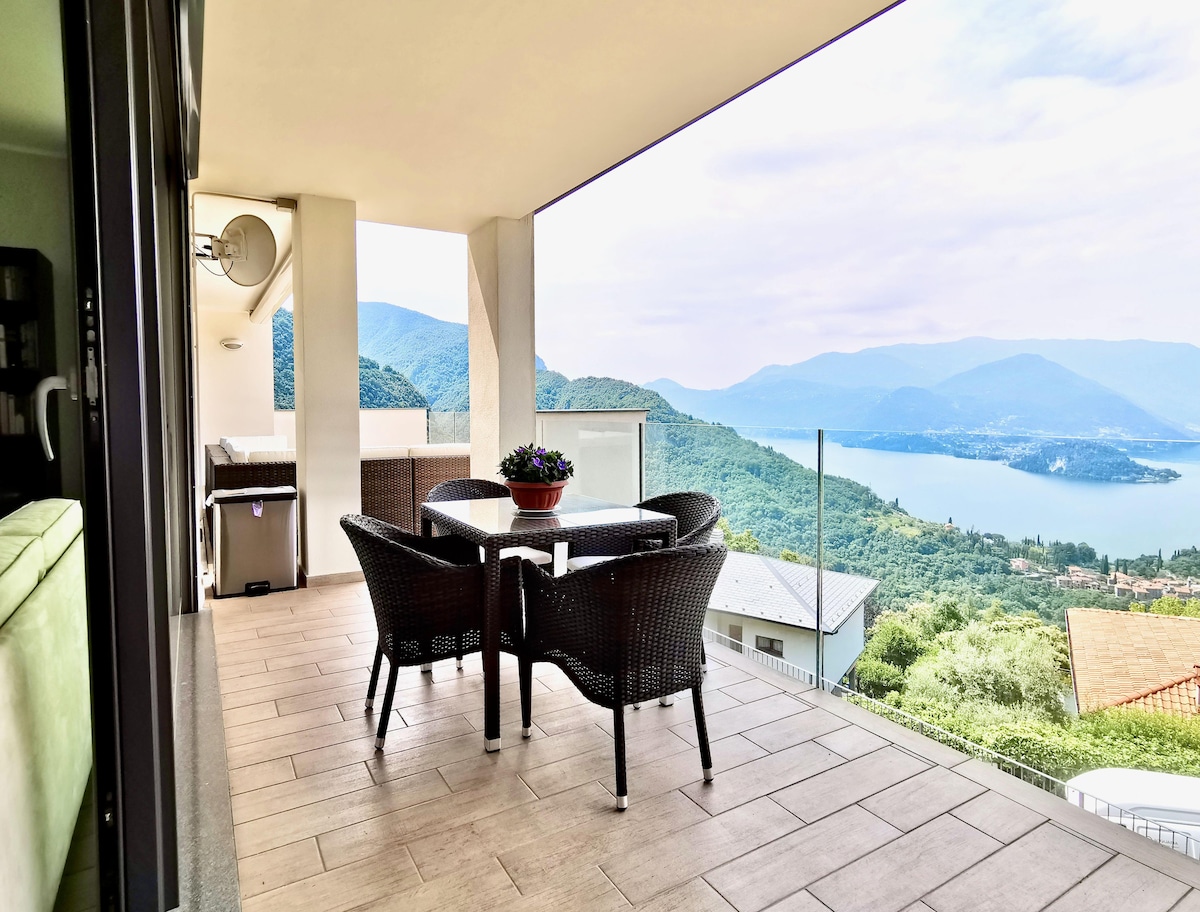 A spacious terrace features a dining table surrounded by chairs, offering a scenic view of rolling hills and a body of water in the distance. A potted plant is positioned at the center of the table, enhancing the outdoor dining experience.