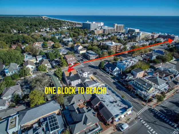 An aerial view showcases the proximity of the beach to the surrounding neighborhood. A clear road leads from the building towards the shoreline, with ocean waves visible in the distance. Various residential structures and businesses fill the streets, emphasizing a vibrant community just steps from the sand.